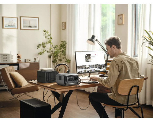 Un bărbat lucrează la un computer cu o centrală electrică portabilă, un router și un dispozitiv de stocare în rețea pe o masă într-o cameră luminoasă.