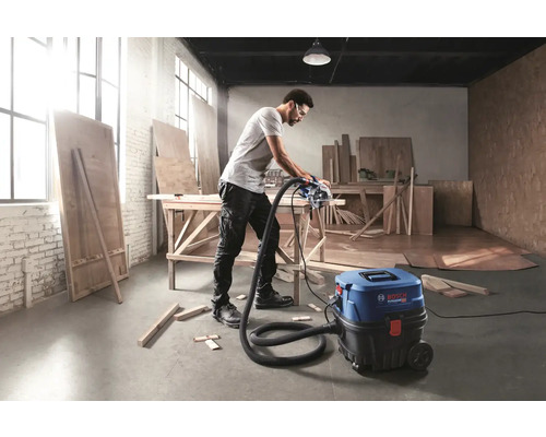 Un bărbat taie o scândură cu un ferăstrău circular conectat la un aspirator Bosch într-un atelier.