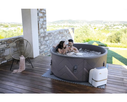 Scenă cu un cuplu relaxându-se într-un jacuzzi gonflabil pe o terasă de lemn. În fundal se vede o zonă rezidențială și dealuri.