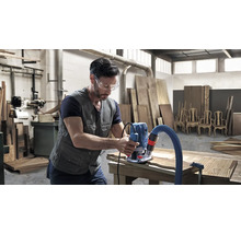 Un bărbat cu ochelari de protecție prelucrează lemn cu o freză superioară într-un atelier.