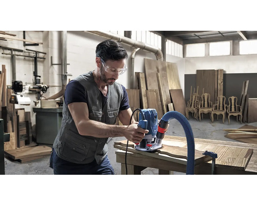 Un bărbat cu ochelari de protecție prelucrează lemn cu o freză superioară într-un atelier.