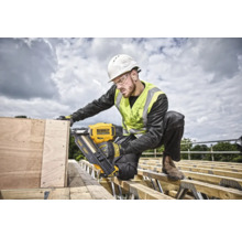 Un muncitor în construcții, purtând cască de protecție, vestă de siguranță și ochelari de protecție, folosește un pistol de bătut cuie cu acumulator DeWalt pe un șantier.