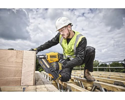 Un muncitor în construcții, purtând cască de protecție, vestă de siguranță și ochelari de protecție, folosește un pistol de bătut cuie cu acumulator DeWalt pe un șantier.