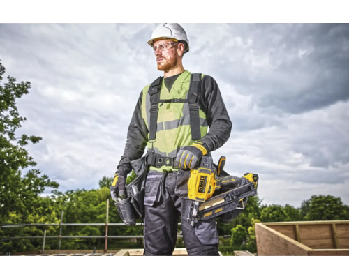 Un muncitor în construcții cu cască de protecție, vestă de siguranță, ochelari de protecție și curea pentru scule poartă un aparat de bătut cuie DeWalt.