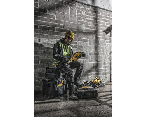 Un meșter cu cască de protecție și vestă de siguranță stă așezat pe cutii negre de scule și ține în mână o mașină de găurit.