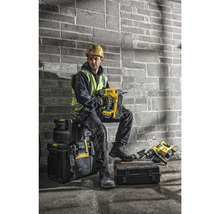 Un meșter cu cască de protecție și vestă reflectorizantă stă așezat pe cutii de scule și ține un pistol de bătut cuie galben marca DeWalt.