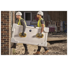 Doi muncitori în construcții care transportă o cădiță de duș folosind ventuze pe un șantier.