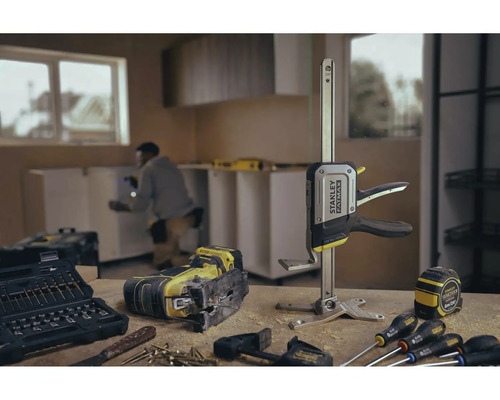 Banc de lucru cu ferăstrău vertical, trusă de scule, clemă cu arc, ruletă și șurubelnițe în fața unei încăperi cu chicinetă.