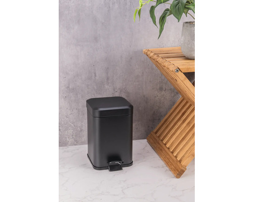 Coș de gunoi negru cu pedală din metal lângă un taburet din lemn cu o plantă