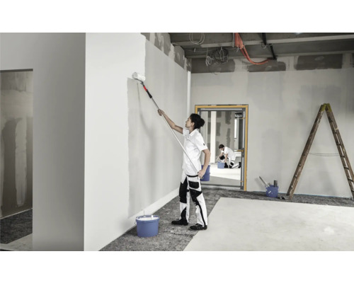 O persoană vopsește un perete într-o cameră cu o rolă de vopsea pe un mâner telescopic. O altă persoană și o scară sunt vizibile în fundal.