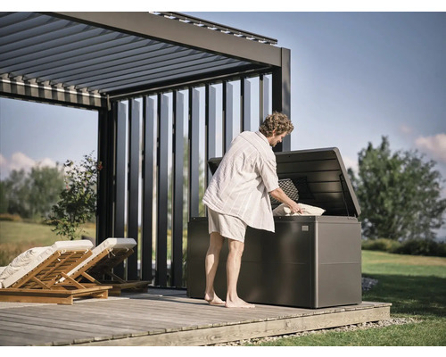 Un bărbat umple o cutie de grădină cu perne sub o pergolă pe o terasă de lemn.