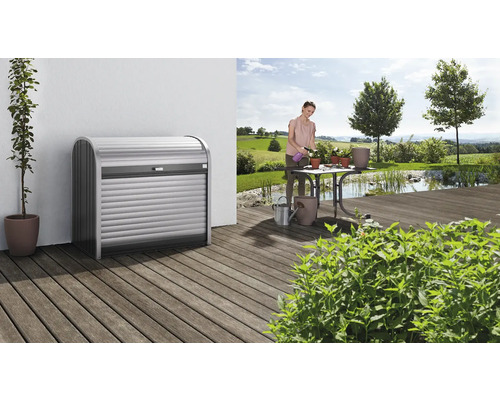 Mască tomberon Biohort StoreMax 120 metal 117x73x109 cm argintiu/gri închis Scenă cu un design de coș de gunoi pe o terasă de lemn și o femeie udând flori în grădină