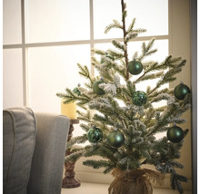 Brad artificial mic, decorativ, cu globuri verzi și sac de iută pe o masă în fața unei ferestre.