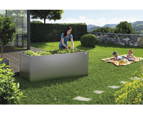 Grădină cu jardinieră metalică, plantată cu legume, în timp ce o femeie recoltează și copiii se joacă.