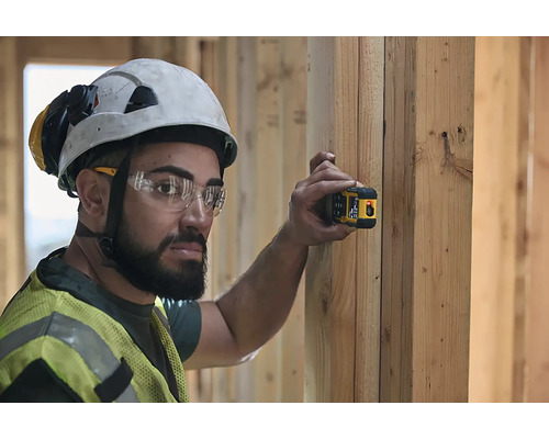 Un muncitor în construcții cu cască de protecție și vestă de siguranță folosește un dispozitiv de măsurare cu laser pe un perete de lemn.