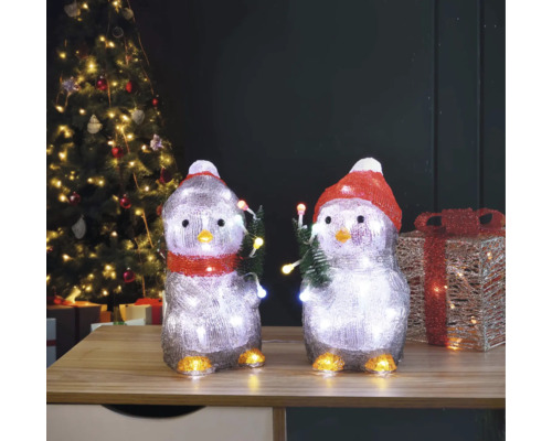 Două figurine de pinguin cu iluminare și motiv de Crăciun pe o masă în fața unui brad de Crăciun.