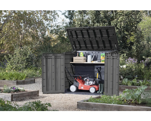 Dulap de grădină cu capac și ușă deschise în grădină, cu mașină de tuns iarba și unelte de grădinărit