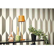 Scenă decorativă cu lampă, lumânări și plantă la ghiveci pe un raft în fața unui tapet geometric.