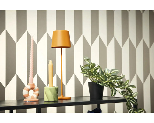 Scenă decorativă cu lampă, lumânări și plantă la ghiveci pe un raft în fața unui tapet geometric.