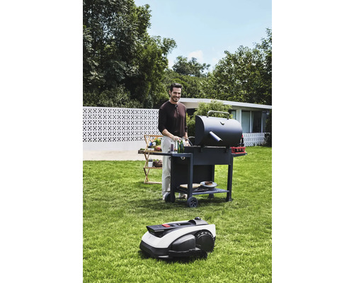 Bărbat care face grătar în grădină cu un grătar și un robot de tuns iarba.