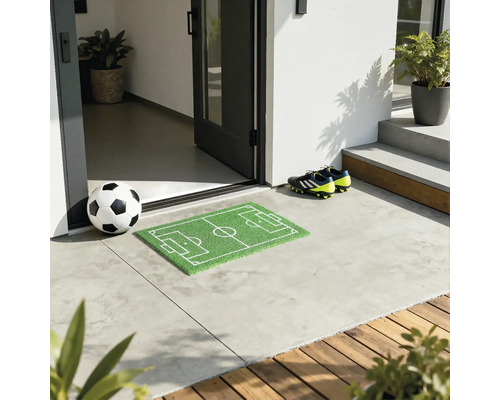 Mingie de fotbal în fața unei uși de intrare cu covoraș de fotbal și ghete de fotbal.