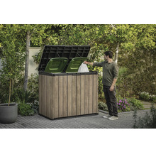 Box pentru pubele din lemn cu două pubele verzi și o persoană care aruncă un sac de gunoi înăuntru