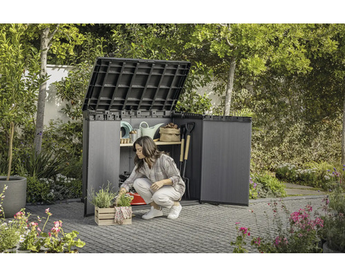 Grădină cu o femeie îngenuncheată în fața unui dulap de grădină deschis în care sunt depozitate unelte de grădină.