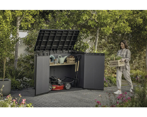 Scenă de grădină cu o femeie care poartă o ladă cu plante lângă o magazie de scule plină cu unelte de grădină și o mașină de tuns iarba