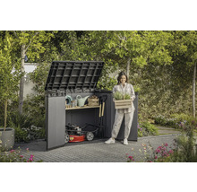 Scenă de grădină cu magazie de scule, unelte de grădinărit și o femeie care ține o ladă cu plante.