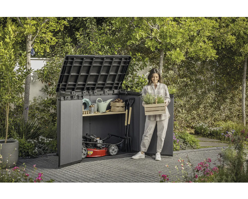 Scenă de grădină cu magazie de scule, unelte de grădinărit și o femeie care ține o ladă cu plante.