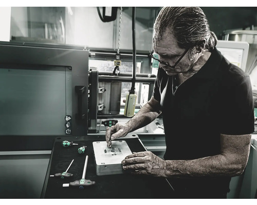 Un bărbat lucrează la un banc de lucru cu unelte și o formă metalică într-un atelier.