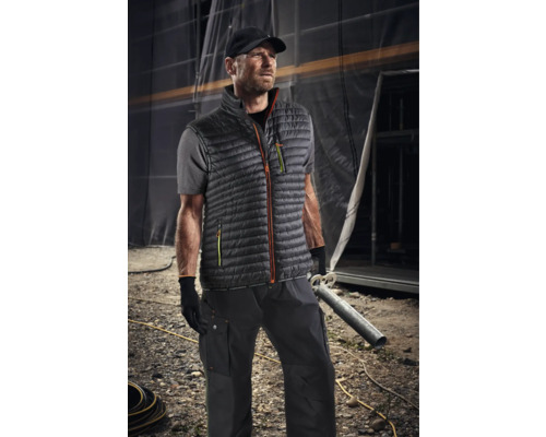 Bărbat purtând haine de lucru cu vestă, tricou, pantaloni și șapcă în fața unui șantier