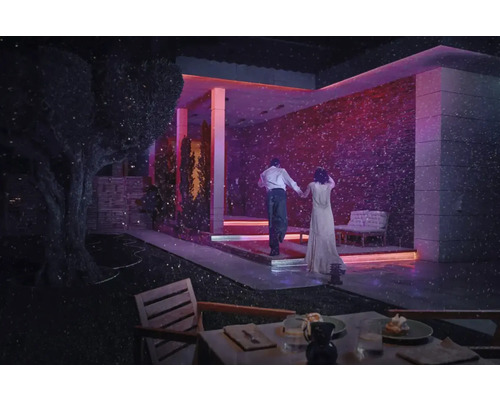 Scenă cu un cuplu plimbându-se de mână în amurg în fața unei case moderne iluminate, cu o masă aranjată în prim plan.