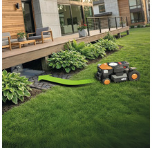 Un robot de tuns iarba se deplasează pe o zonă verde către stația sa de încărcare sub o terasă de lemn.
