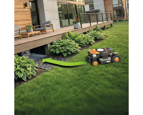 Un robot de tuns iarba se deplasează pe o zonă verde către stația sa de încărcare sub o terasă de lemn.