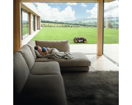 Băiat doarme pe canapea în timp ce robotul de tuns iarba lucrează în grădină