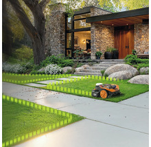 O mașină de tuns iarba robotizată pe un gazon în fața unei case cu fațadă de piatră și un copac mare.