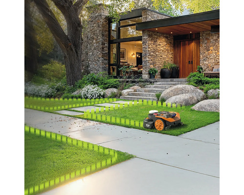 O mașină de tuns iarba robotizată pe un gazon în fața unei case cu fațadă de piatră și un copac mare.