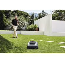 Un robot de tuns iarba pe un gazon, cu un bărbat care se plimbă cu o băutură în fundal.