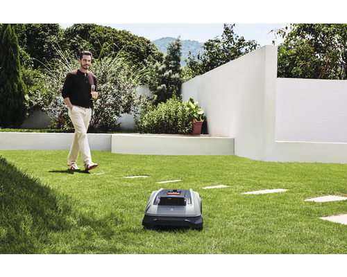 Un robot de tuns iarba pe un gazon, cu un bărbat care se plimbă cu o băutură în fundal.