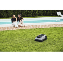 Robot de tuns iarba pe gazon în fața unui cuplu așezat lângă o piscină.