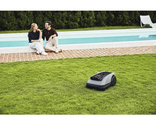 Robot de tuns iarba pe gazon în fața unui cuplu așezat lângă o piscină.