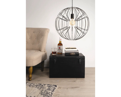 Scenă cu un fotoliu, un cufăr negru, cărți, o băutură și o lampă suspendată cu cadru metalic.