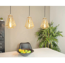 Trei lămpi suspendate aurii deasupra unei mese cu un bol de fructe și o plantă în fundal.