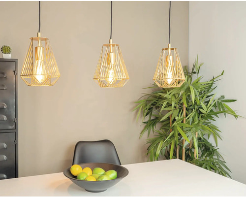Trei lămpi suspendate aurii deasupra unei mese cu un bol de fructe și o plantă în fundal.