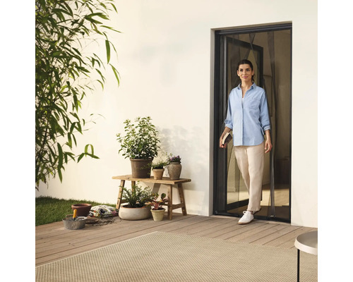 Femeie intrând pe terasă printr-o ușă cu plasă de insecte, înconjurată de plante și accesorii de grădină.