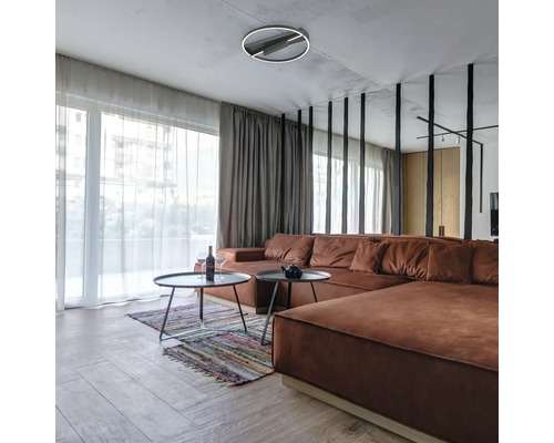Living modern cu o canapea de colț maro, două măsuțe de cafea rotunde, un covor colorat și o plafonieră LED circulară.