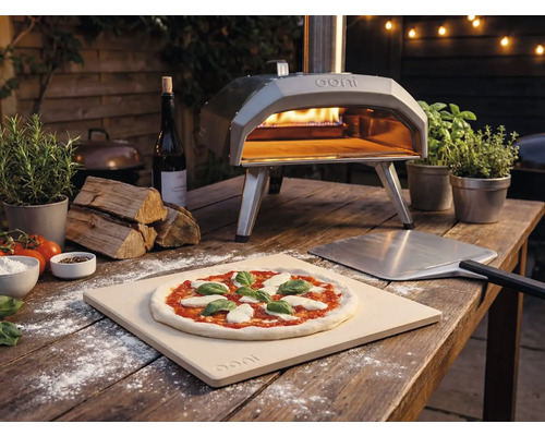 Imagine exterioară cu un cuptor de pizza Ooni cu pizza, paletă de pizza și ingrediente pe o masă de lemn.