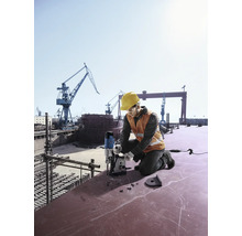 Un muncitor în construcții cu cască de protecție și vestă de siguranță forează cu o mașină de găurit magnetică pe un șantier naval.