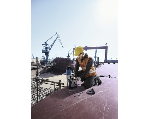 Un muncitor în construcții cu cască de protecție și vestă de siguranță forează cu o mașină de găurit magnetică pe un șantier naval.